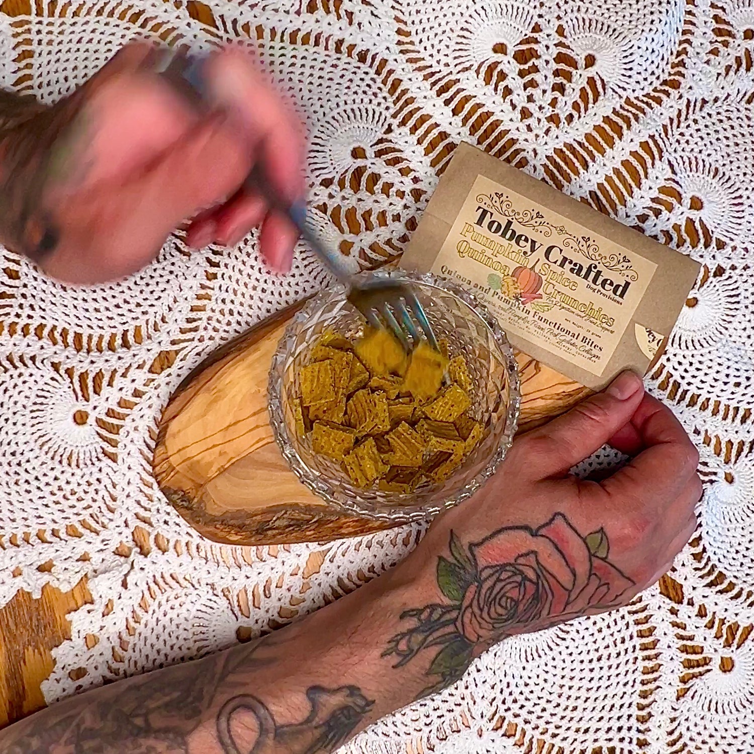 Person holding a bowl of dog treats with a 'Tobey Crafted' packaging on a lace tablecloth.