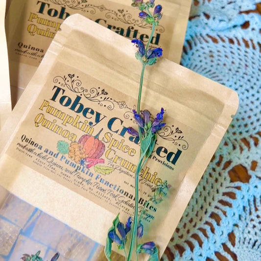 Tobey Crafted Pumpkin Spice Quinoa Crunchie packets with a sprig of sage flower on a crochet doily background