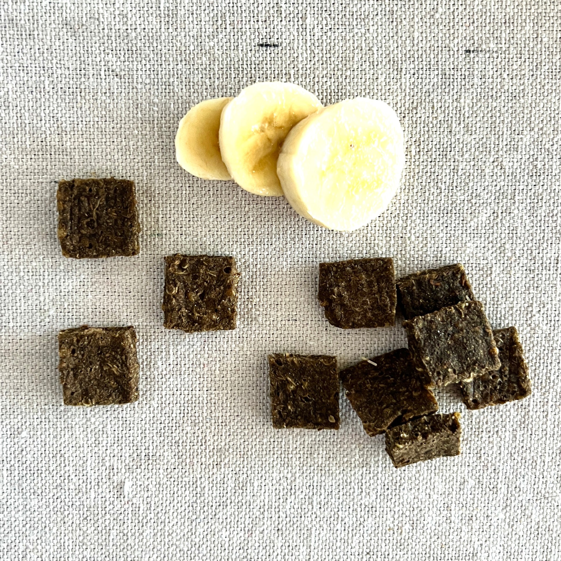 Brown square-shaped banana balm tiles and a slice of banana on a textured gray surface