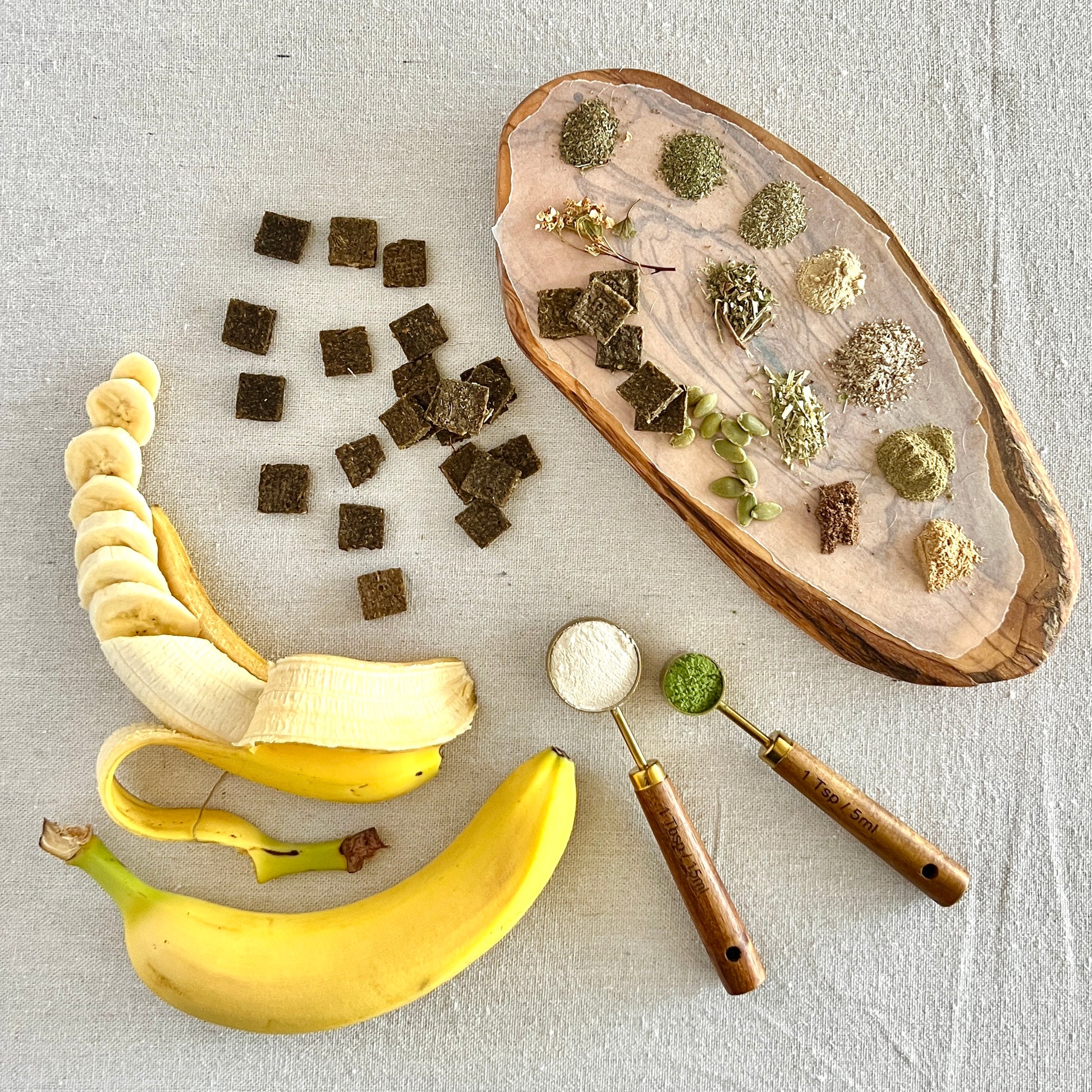 Bananas, a wooden cutting board with  herbs, crunch n’ care tiles, and a wooden spoon on a light gray background.