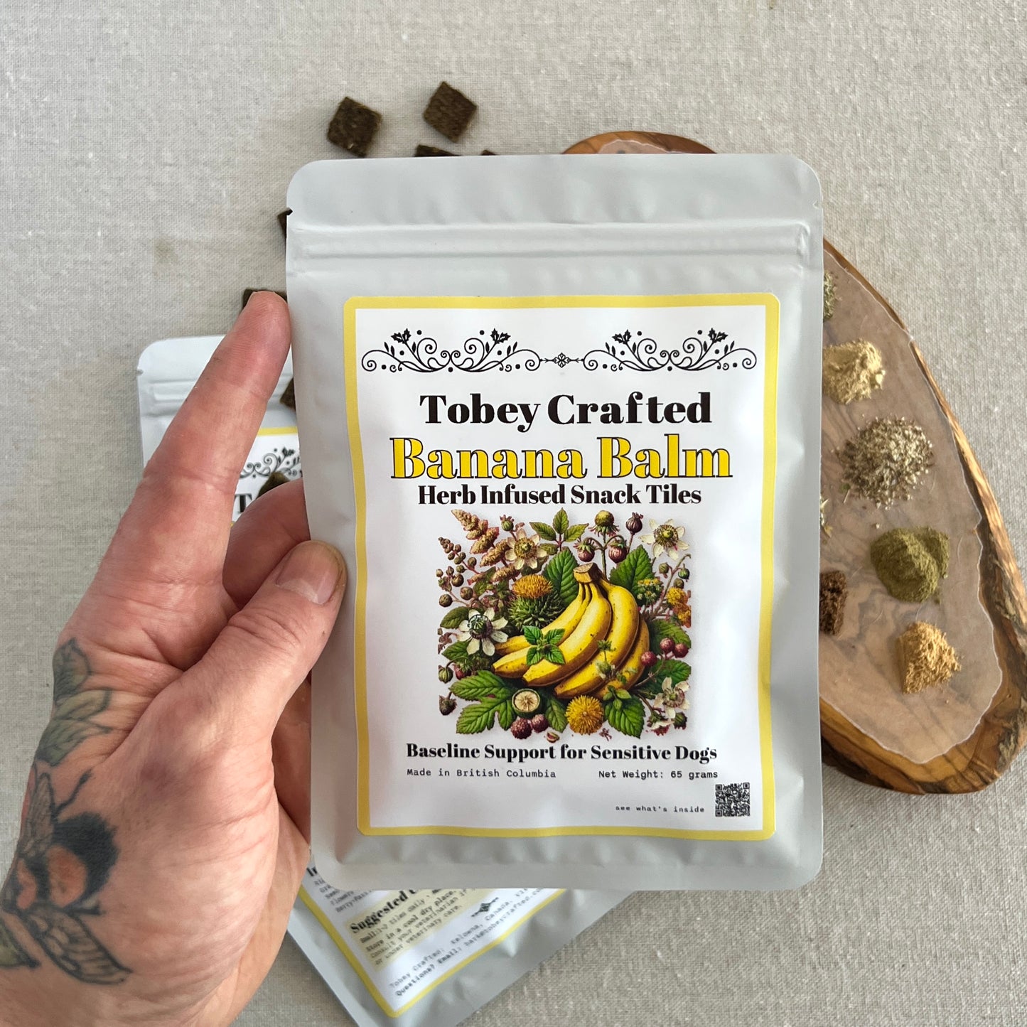Hand holding a package of Tobey Crafted Banana Balm Herb Infused Snack Tiles on a neutral background.