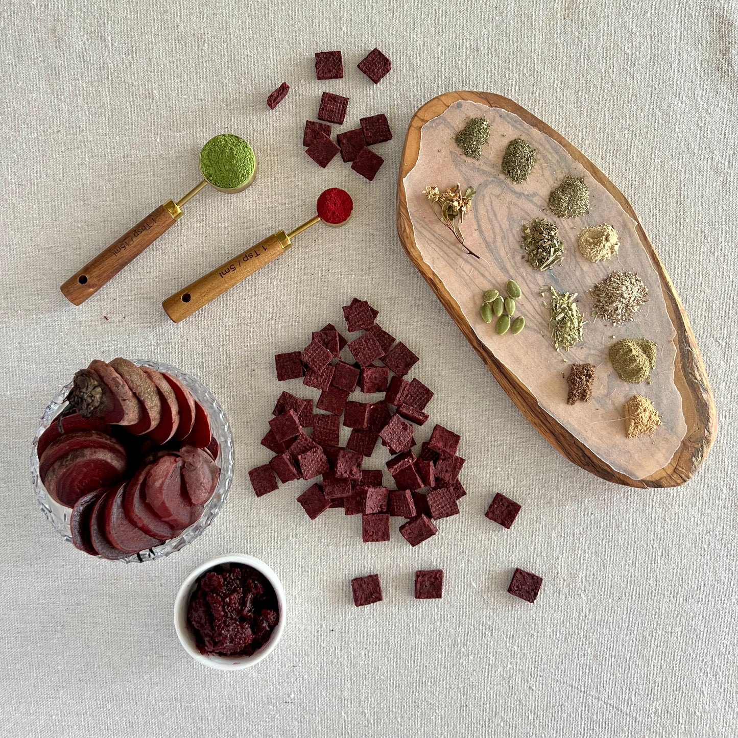 Assorted dried herbs and cooked beets on a textured surface with a wooden scoop and small containers.
