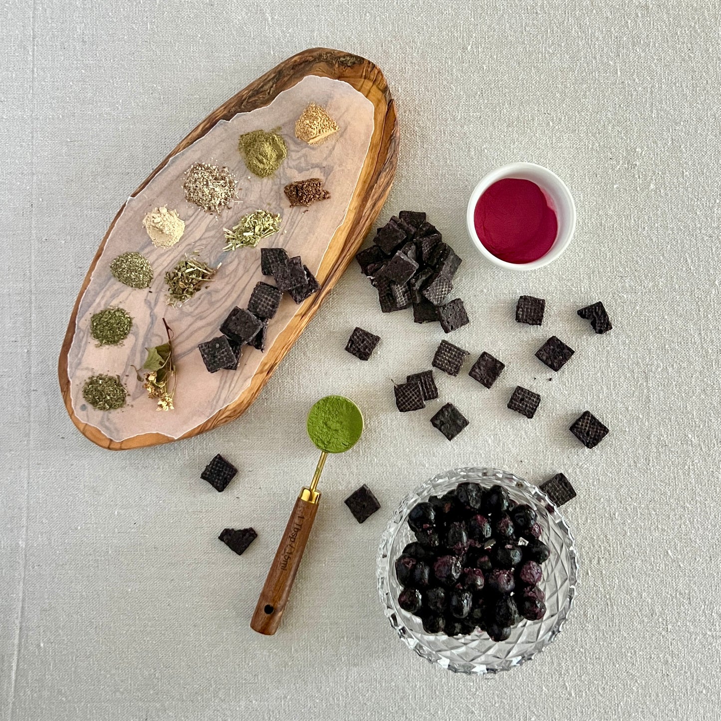 Wooden tray with dried herbs, Blueberry Ease tiles, blueberries, a small bowl of blueberry powder on a light gray background.