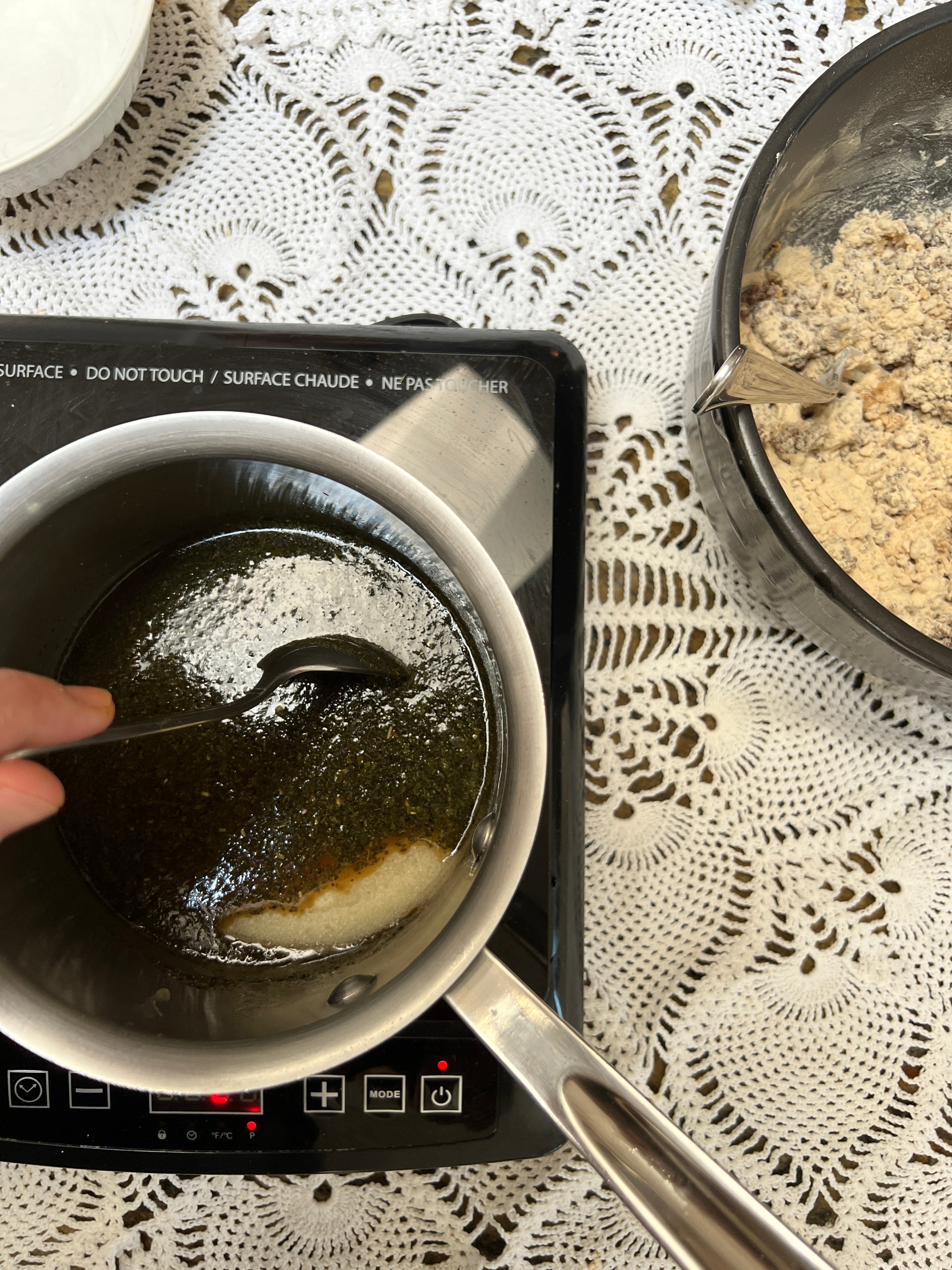 Stirring dark liquid in a pot on a small stove with a lace tablecloth in the background.